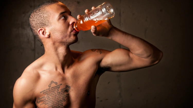 A topless man with a tattoo on his chest and shoulder chugs a sports drink from a clear bottle in a dimly lit studio.