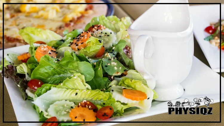 A chopped salad toped with tomatoes, carrots and sesame seeds is sitting on a white plate with a small white pouring saucer beside it and next to a cheese pizza.
