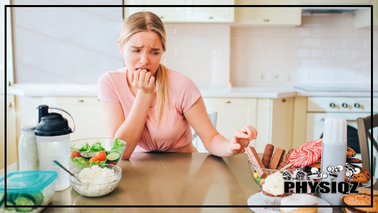 A woman in a short sleeve ping shirt is thinking of different ways to hack hunger while fasting because she's sitting at her beige kitchen table and reaching towards a chocolate candy bar sitting in a glass bowl as she makes a face of desperation since she feels like she's starving.