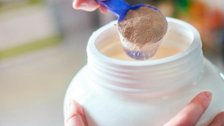 A person is holding a white container of protein powder and using a blue scooper to get some powder, and it appears that the powder is in chocolate flavor.