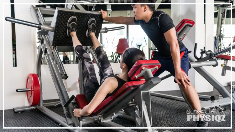 On the right is a man in blue shorts and he is helping out a woman who is lying horizontally on a red pad that's attached to a leg press machine and her legs are straight since she just pushed the weight up. 