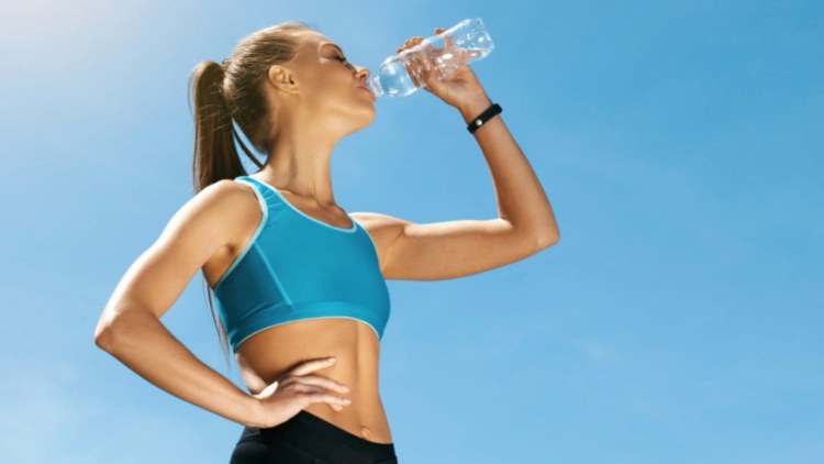 A woman wearing a blue sports bra and black bottoms is drinking a water from a clear water bottle in an outdoor setting with a view of clear blue sky in the background.
