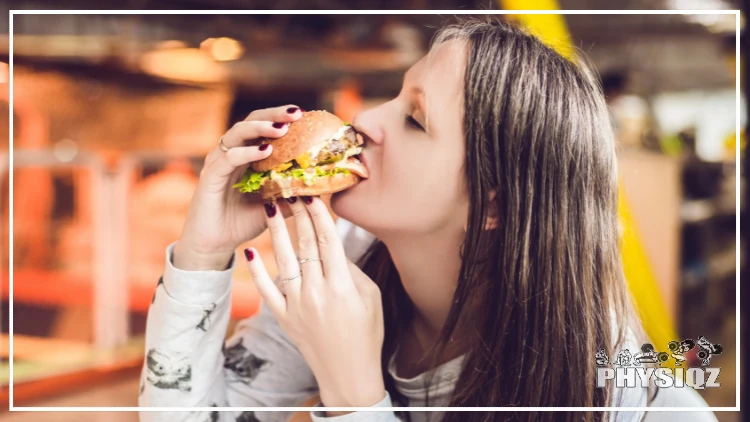 A white woman with straight brunette hair has her nails painted dark red, wearing a white long sleeve shirt and is taking a bite from a large burger that has beef, lettuce, onions and cheese on it as she sits on the patio of a restaurant. 