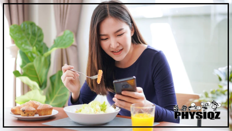 An Asian woman is holding a fork that has a carrot on it in her right hand and her cell phone in her left hand while sitting at a table with blueberry muffins, lettuce, and orange juice on it. 