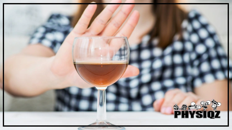 A wine glass full of alcohol is sitting in a white table and a woman with red hair and white and black dotted shirt is in the background with her hand in the "stop" position since she's saying no to drinking. 