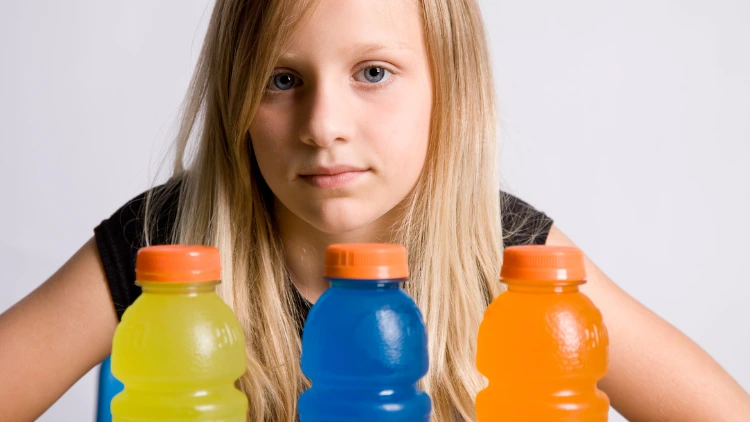 A blonde young girl wearing a black blouse looks at the camera while there are three clear bottles of sports drink with orange cap in front of her in different colors such as yellow, blue and orange.