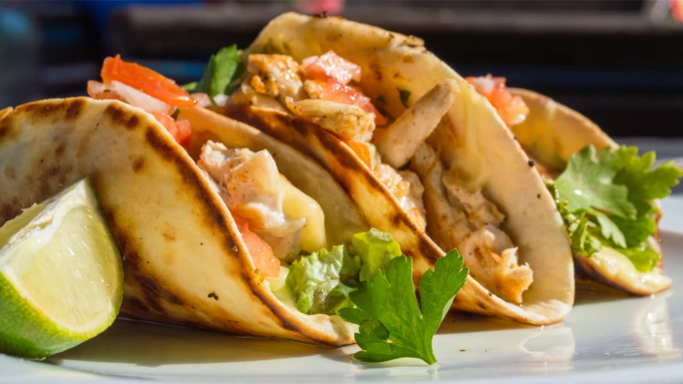 A plate of three chicken tacos with a slice of lime, each taco is filled with seasoned chicken, lettuce, and chopped tomatoes, and is served on a warm tortilla, the tacos are arranged on a white plate.