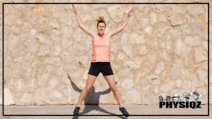 A woman with her hands in the air and her legs spread since she's doing one of a jumping back after committing to 100 jumping jacks a day and there's a wall made out of rocks behind her.