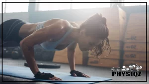 A woman with her hair pulled back into a ponytail is doing the 500 pushups a day challenge with her gloved hands on a blue mat in a gym while mid-pushup, wearing a light blue tank top and in the background are weight plates and box jumps stacked on the floor.