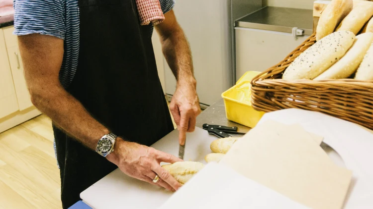 A man with a silver watch and a ring wearing a striped blue and white t-shirt and black apron is making a sandwich in a store kitchen counter using a bread knife on a white chopping board, and on the counter, there are basket filled with sandwich buns.