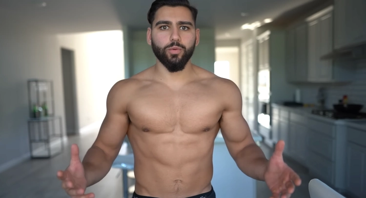 A man with a beard flexing his muscles to show his chest is a little bigger from doing push ups, and the background is in a kitchen setting with countertops and appliances visible in the frame.