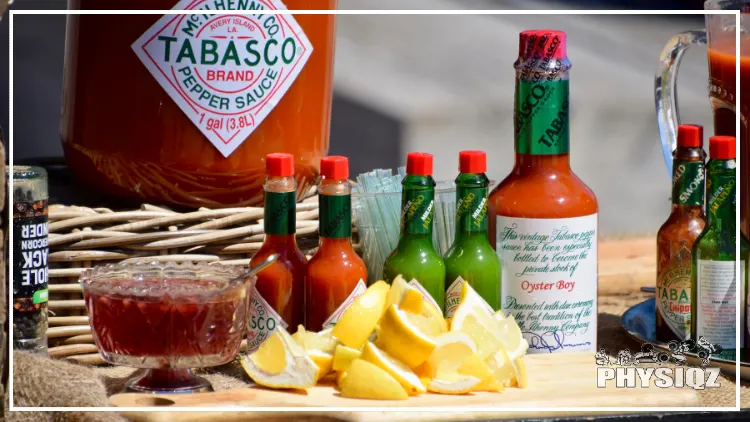 A large Tabasco hot sauce bottle in the background with smaller assorted bottles that are green and red in the foreground as well as a wooden cutting board with lemons even further to the front. 