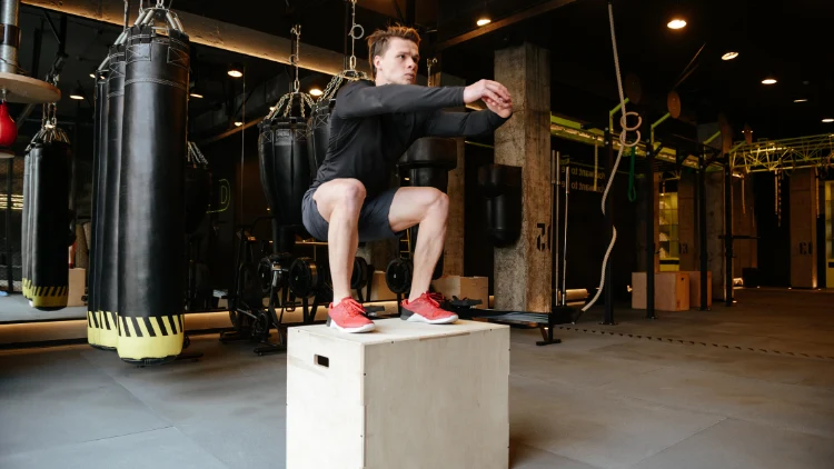An athletic young man wearing a black sweater and shorts, and red shoes is jumping on a plyo box in a gym with lines of black punching bags in the background.