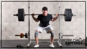 Inside a room that has a power rack, rubber mats, and cinderblock walls a man in white pants, a black shirt, white shoes and grey knee sleeves is executing a back squat workout with 245 pounds on the bar as he descends into the bottom of the squat.