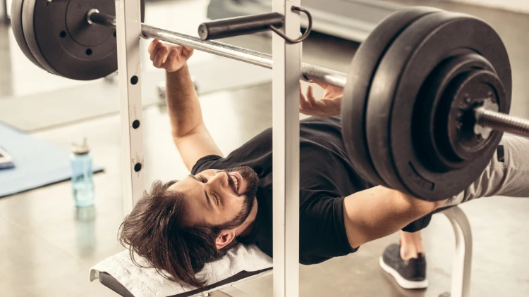 A bearded guy wearing a black t-shirt, grey shorts and black shoes is on a bench about to grab the barbell with two weighted plates as he prepares to perform a bench press exercise.