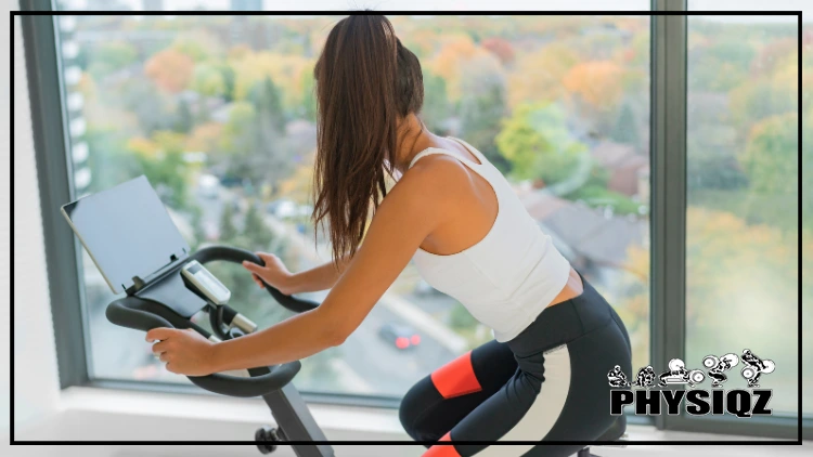 A slender woman with her hair tied back, dressed in a white tank top and black leggings, rides a Peloton bike while watching a beginner class led by top Peloton instructors in a studio with large glass windows.