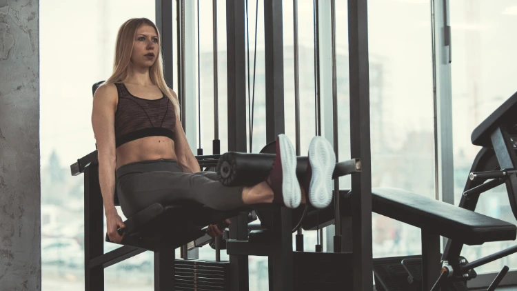 A blonde woman wearing a black sports bra, grey leggings and red-ish shoes is performing a seated leg curls using the machine in a gym with huge glass window in the background.