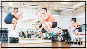 Five people are in the middle of a jump squat which is a box jump alternative commonly used, and the people are attending a gym workout class where there's an instructor, wooden floors, and weights in the background.