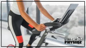 A tan woman in a white sleeveless shirt and black pants is wondering can you lose weight with Peloton while pedaling indoor on a stationary bike that's in a white room and has a forest landscape seen out the window.