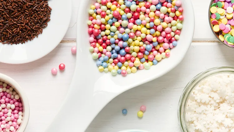 A white pan filled with assorted colorful candy beads such as blue, yellow, red, and pink, beside it, on the left, is a white plate of a chocolate sprinkles, below it is a white bowl of pink bead candies, and on the right, is another assorted colorful candies, and below it is a bowl of white candies.
