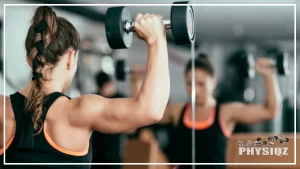 A white woman with braided brunette hair is doing one of the many compound shoulder exercises or a dumbbell over head press in front of a mirror that shows her blurry reflection and has on a black tank top as she holds the black and silver dumbbells' up above her head.