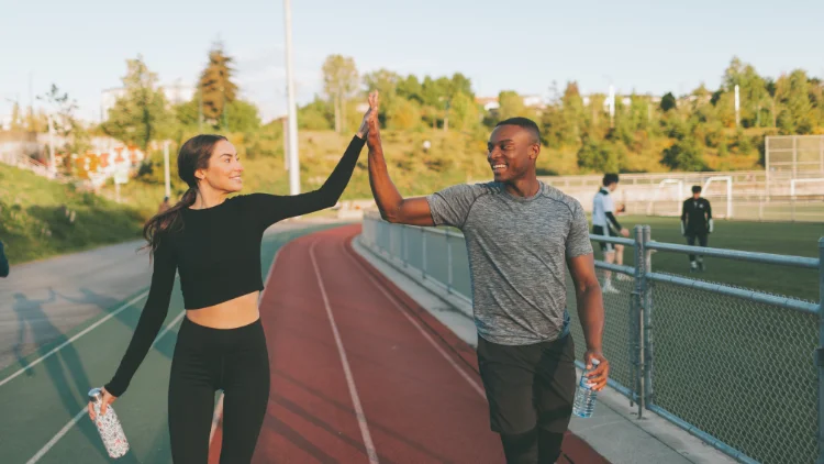 On the left is a white woman with brown hair and black attire on and on the right is a black man in a grey shirt and they're both high fiving while holding water bottles and there's a track and field in the background. 