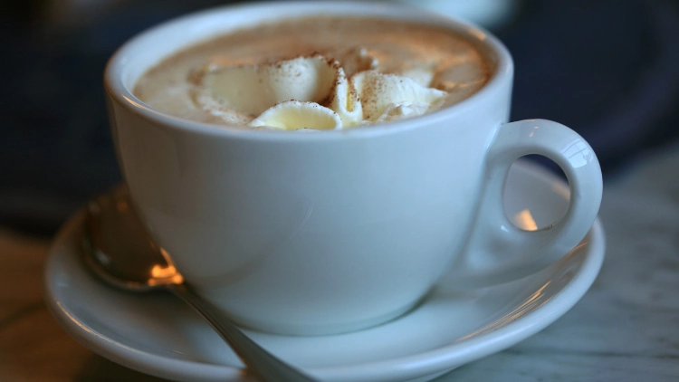 A white cup filled with frothy hot chocolate, served on a small dish plate with a spoon,