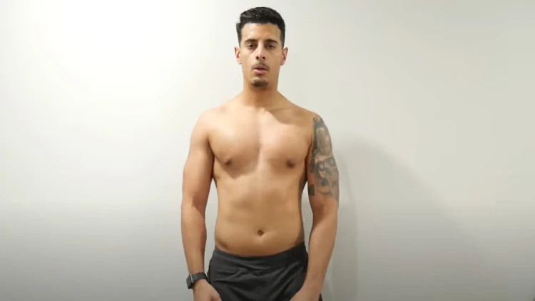 A man with tattoos on his left arm and a watch on his right arm is wearing a black short and showing off his body's workout progress.