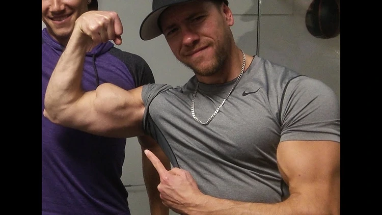 The man in the gray shirt has on silver necklace and is flexing his right bicep that has an extremely high insertion point, while the man on his right is wearing a purple shirt and smiling. 