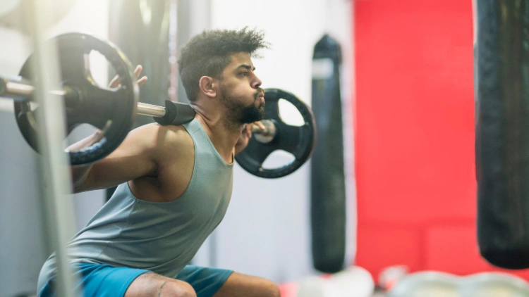 A fit man with beard and slightly curly hair, wearing a cyan tank top and blue short is performing a barbell back squat using a barbell with only one weighted plate in a gym with red portion on the wall in the background.
