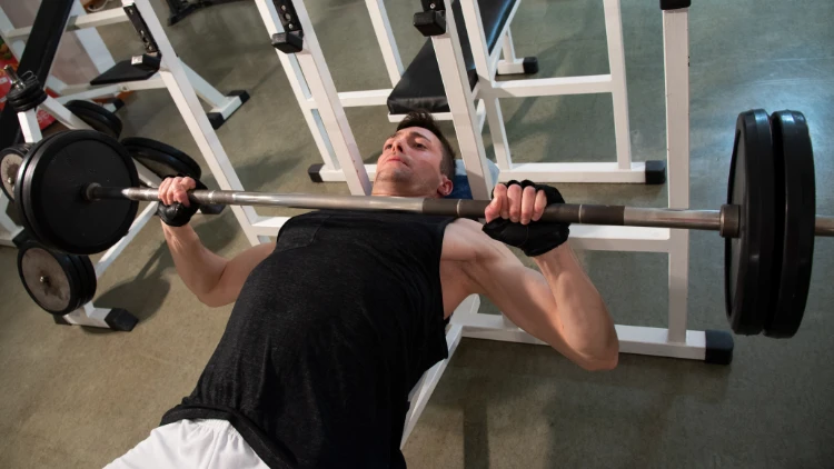 A fit young man wearing a black tank top and white short is performing a bench press using a barbell with two weighted plates in a gym with concrete floor.