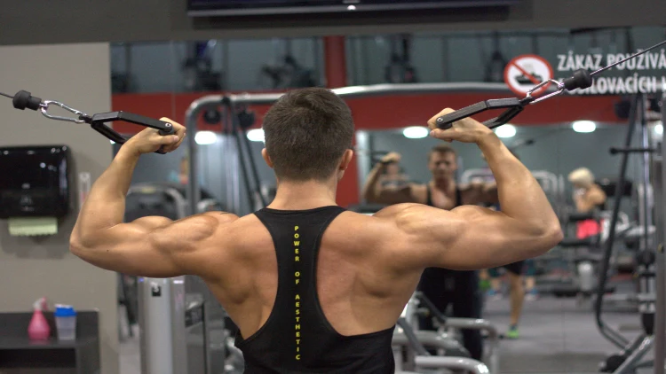 A man wearing black tank top doing an exercise using an equipment in a gym while looking at his reflection.