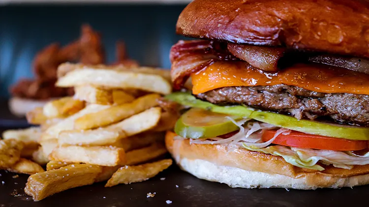 A closeup photo of a burger stuffed with bacon, lettuce, tomato and cheese, and on the side is a serving of fries and displayed on a shiny black surface.