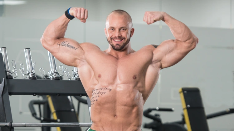 A jacked white guy is doing a front double bi pose that shows his low tricep insertions and he's wearing a watch on his right wrist, he's got a tattoo on his right bicep and his rib area.