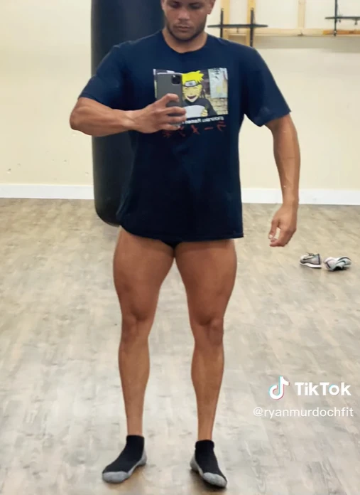 A guy wearing a blue shirt, taking a mirror picture showing his legs with super small calves in comparison to the rest of his body, especially his quads. 