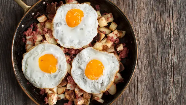 Hash brown breakfast with three sunny side up eggs on top served in a skillet and placed on top of a wooden surface.