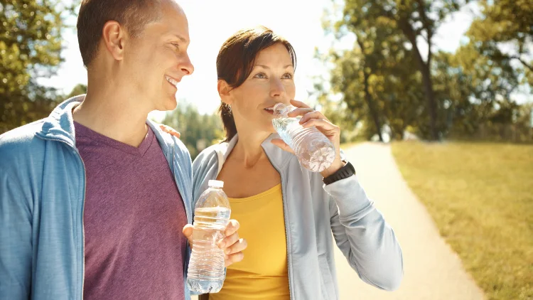 A healthy couple outdoors, the woman is wearing a yellow tank top with a white jacket, and the man is wearing a red-ish shirt with blue jacket, both of them are holding a clear water bottle.