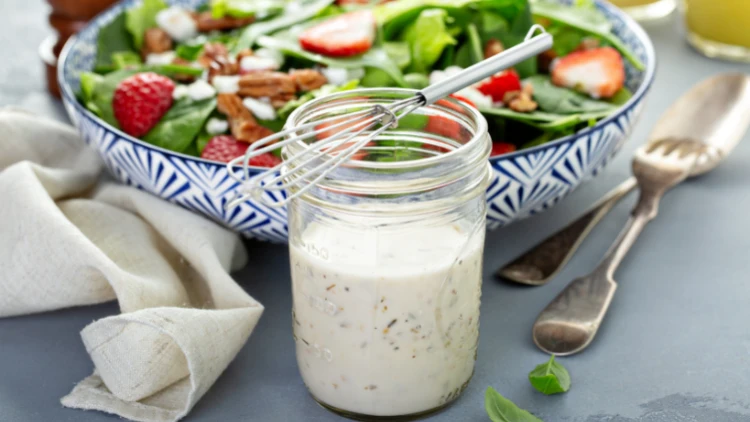 A jar of homemade ranch dressing with a whisk sitting on top of the jar's lid, the jar is filled with a creamy white dressing with specks of herbs visible throughout, in the background, a bowl of fresh salad with lettuce, tomatoes, carrots, and cucumbers is visible, ready to be dressed.