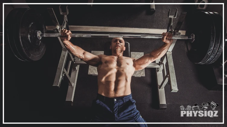 A topless muscular man wearing a dark blue short is performing a bench press of 3 weighted plates while wondering how many people can bench 315 pounds like him in a dimly lit gym, with dark flooring.