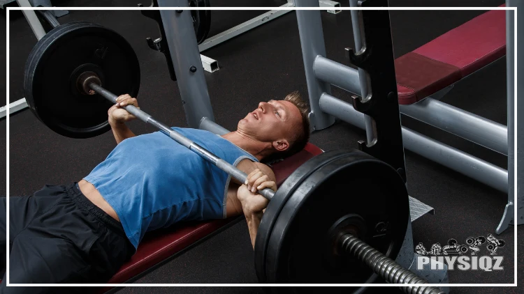 A guy in blue tank top and black short who's been asking himself how many reps of 225 to bench 315 while performing bench press exercise using a barbell with two weighted plates in a gym with dark-colored floor.