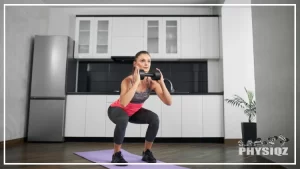 A woman in grey shorts, black shoes, and a pink top is implementing tips on how to gain weight in legs for skinny girls by doing a dumbbell squat inside her kitchen that has dark wooden floors, white cabinets, stainless steel fridge, and a house plant in the corner.