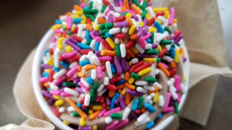 Ice cream with colorful sprinkles such as pink, violet, green, white, blue, orange and yellow in a small white cup placed on top of a brown napkin.