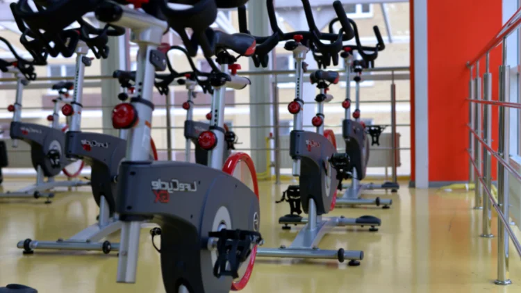 Five indoor stationary bikes that are black, red and grey in color are sitting on a light yellow floor inside a gym studio with silver railings and red walls. 