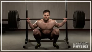 A topless powerlifter, wearing a black short, shoes and socks is doing a deep squat using a barbell with 4 weighted plates and wonders "Is 405 squat good weight, or is it just okay?", in a dimly lit gym with concrete wall and floor.