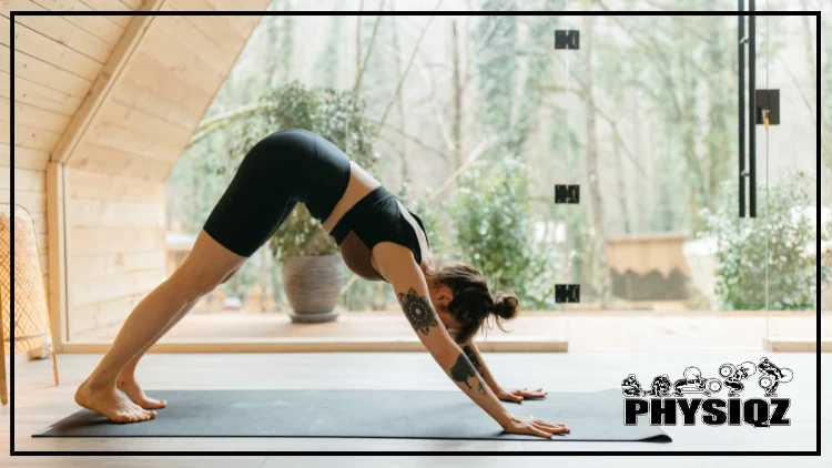 A woman with her hair in a bun is wondering is Pilates good for weight loss while wearing a black and brown tank top and black sport shorts while bending forward on a cool grey tone yoga mat and doing a downwards facing dog and there are plants and trees in the background.