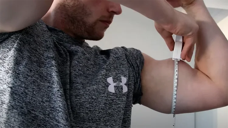 A man in a grey Under Amour shirt is measuring the circumference of his left bicep with a white measuring tape. 