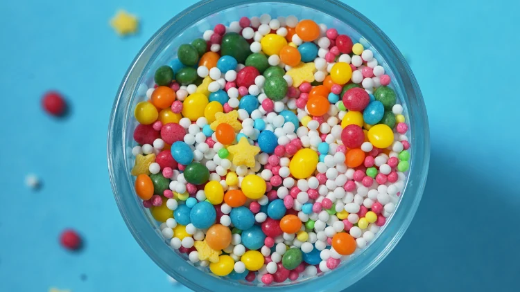 Assorted colorful candies and sprinkles such as yellow, blue, orange, pink, red and white in a clear glass cup placed on top of a blue surface with some candy spills.