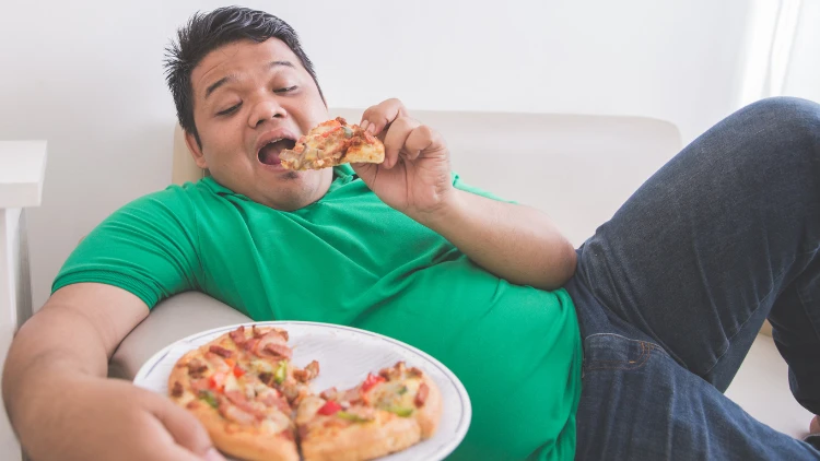 An overweight man in a green shirt, and dark denim pants on a beige colored couch eating a plate of pizza that has onions, peppers, ham and pepperoni on it. 