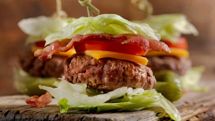 A burger made with beef patty, cheese, tomato and bacon wrapped in lettuce, behind it are the same two lettuce wraps and displayed on a wooden board.