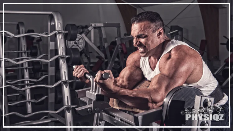 A tan man in a white tank top is doing machine preacher curls on a grey machine and inside a gym with unidentifiable equipment in the background.